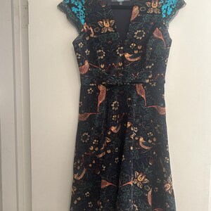 K&ouml;nnte beinhalten: Dunkelblaues Kleid mit V-Ausschnitt und Fl&uuml;gel&auml;rmeln. Das Kleid hat ein Blumen- und Vogelmuster in Orange-, Gold- und Gr&uuml;nt&ouml;nen. Die Fl&uuml;gel&auml;rmel haben ein t&uuml;rkisfarbenes Paillettendetail. Das Kleid h&auml;ngt an einem schwarzen Kleiderb&uuml;gel.