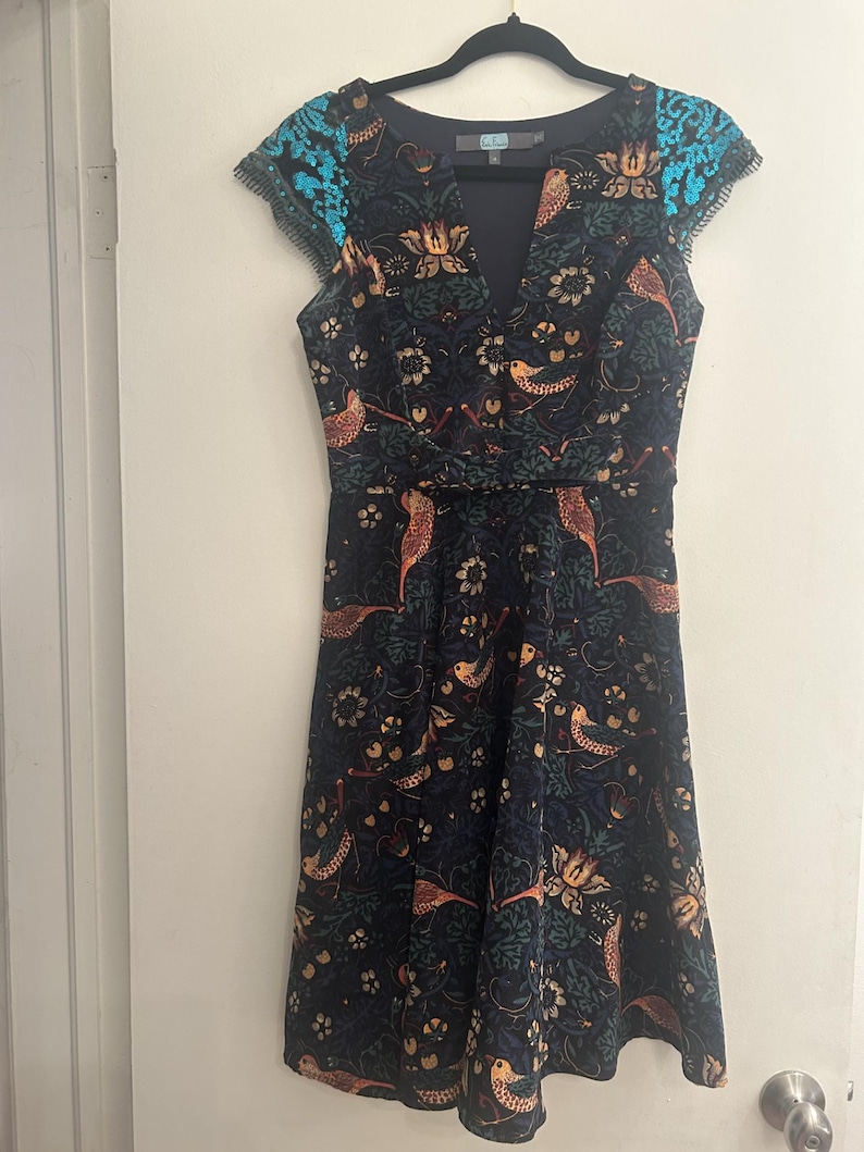 K&ouml;nnte beinhalten: Dunkelblaues Kleid mit V-Ausschnitt und Fl&uuml;gel&auml;rmeln. Das Kleid hat ein Blumen- und Vogelmuster in Orange-, Gold- und Gr&uuml;nt&ouml;nen. Die Fl&uuml;gel&auml;rmel haben ein t&uuml;rkisfarbenes Paillettendetail. Das Kleid h&auml;ngt an einem schwarzen Kleiderb&uuml;gel.