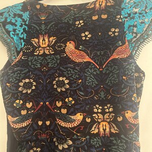K&ouml;nnte beinhalten: Ein &auml;rmelloses Kleid mit einem dunkelblauen Blumen- und Vogelmuster. Das Kleid hat einen runden Ausschnitt und t&uuml;rkisfarbene Pailletten-Details an den Schultern. Der Stoff hat ein detailliertes botanisches Design mit V&ouml;geln.