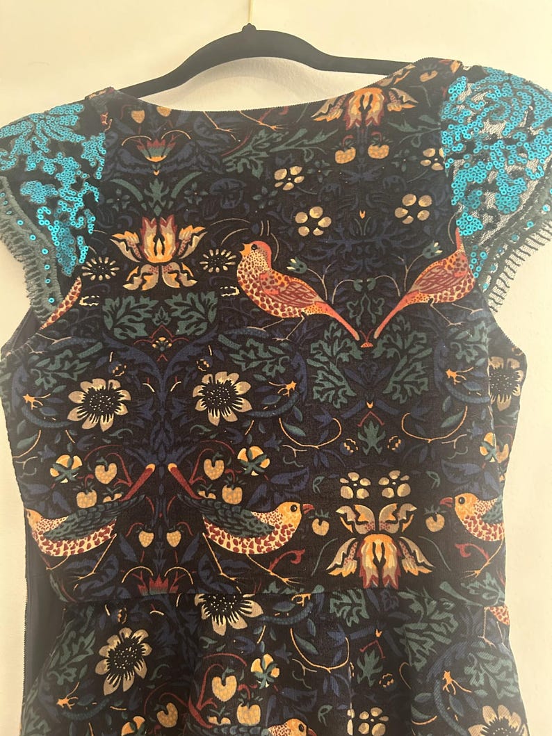 K&ouml;nnte beinhalten: Ein &auml;rmelloses Kleid mit einem dunkelblauen Blumen- und Vogelmuster. Das Kleid hat einen runden Ausschnitt und t&uuml;rkisfarbene Pailletten-Details an den Schultern. Der Stoff hat ein detailliertes botanisches Design mit V&ouml;geln.