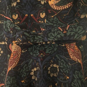 K&ouml;nnte beinhalten: Nahaufnahme eines dunkelblauen Stoffes mit einem sich wiederholenden Muster aus V&ouml;geln, Blumen und Bl&auml;ttern in Orange, Gr&uuml;n und Gold. Der Stoff scheint ein Kleid oder ein Oberteil zu sein, mit einem gerafften Detail in der Taille.