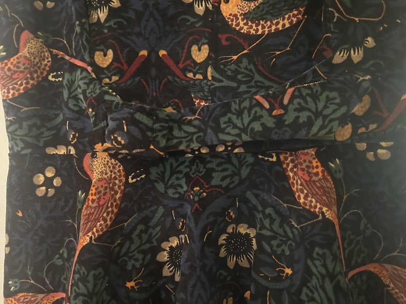 K&ouml;nnte beinhalten: Nahaufnahme eines dunkelblauen Stoffes mit einem sich wiederholenden Muster aus V&ouml;geln, Blumen und Bl&auml;ttern in Orange, Gr&uuml;n und Gold. Der Stoff scheint ein Kleid oder ein Oberteil zu sein, mit einem gerafften Detail in der Taille.