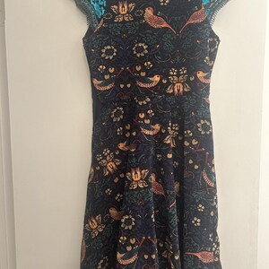 K&ouml;nnte beinhalten: Ein &auml;rmelloses, knielanges Kleid mit dunkelblauem Hintergrund und einem Muster aus V&ouml;geln und floralen Designs in Orange-, Gr&uuml;n- und Goldt&ouml;nen. Die Schultern sind mit t&uuml;rkisfarbenen Pailletten verziert. Das Kleid h&auml;ngt an einem schwarzen Kleiderb&uuml;gel.