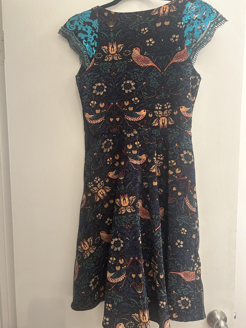 K&ouml;nnte beinhalten: Ein &auml;rmelloses, knielanges Kleid mit dunkelblauem Hintergrund und einem Muster aus V&ouml;geln und floralen Designs in Orange-, Gr&uuml;n- und Goldt&ouml;nen. Die Schultern sind mit t&uuml;rkisfarbenen Pailletten verziert. Das Kleid h&auml;ngt an einem schwarzen Kleiderb&uuml;gel.
