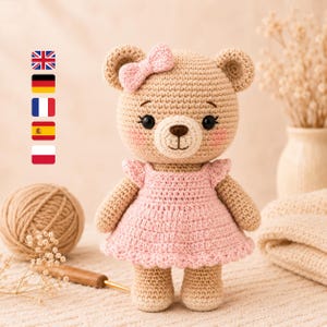 Könnte beinhalten: Gehäkelter Teddybär mit rosa Kleid und Schleife. Der Bär hat eine braune Nase, schwarze Augen und rosige Wangen. Ein Wollknäuel und eine Häkelnadel befinden sich im Hintergrund und deuten auf ein Bastelprojekt hin.