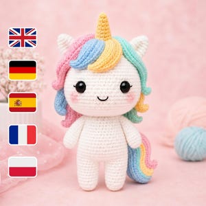 Puede incluir: Una muñeca de unicornio de ganchillo blanco hecha a mano con un cuerno amarillo, melena rosa, azul y amarilla y una cola de arcoíris. El unicornio tiene ojos negros y mejillas rosadas. La muñeca está sobre una superficie rosa.