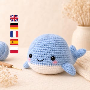 Op de afbeelding: Een gehaakte blauwe walvis met een witte buik, zwarte ogen en roze wangen. Het handgemaakte pluche speelgoed heeft kleine vinnen en een lachende mond. Een bol blauw garen en een haaknaald zijn zichtbaar.