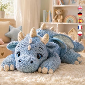 May include: A large, light blue crochet dragon plush toy featuring cream-colored horns, spikes along its back, and claws. The dragon has large, expressive blue eyes with black pupils and detailed stitching, lying in a resting position.
