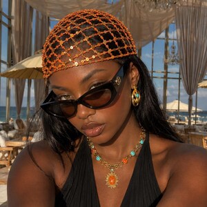 Op de afbeelding: Een oranje kralen hoofdtooi met een netpatroon, zwarte cat-eye zonnebril, goudkleurige oorbellen met turquoise accenten en een goudkleurige ketting met afwisselend turquoise en oranje kralen en een centrale ovale oranje hanger.