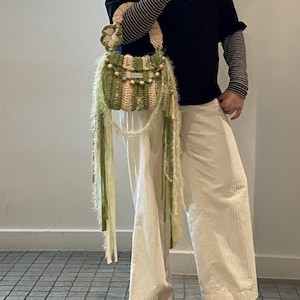 May include: A handmade crochet handbag in shades of green and beige, adorned with beaded accents and long, flowing fringe. The bag features a floral detail on the handle and a striped pattern. The bag is carried by a person wearing a black shirt and cream-colored pants.