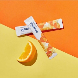 May include: Two white Unicity Balance Orange supplement packets with a geometric orange and yellow circle pattern. Each packet displays the text Unicity Balance Orange and a net weight of 0.25 oz. A fresh orange wedge sits beside the packets.