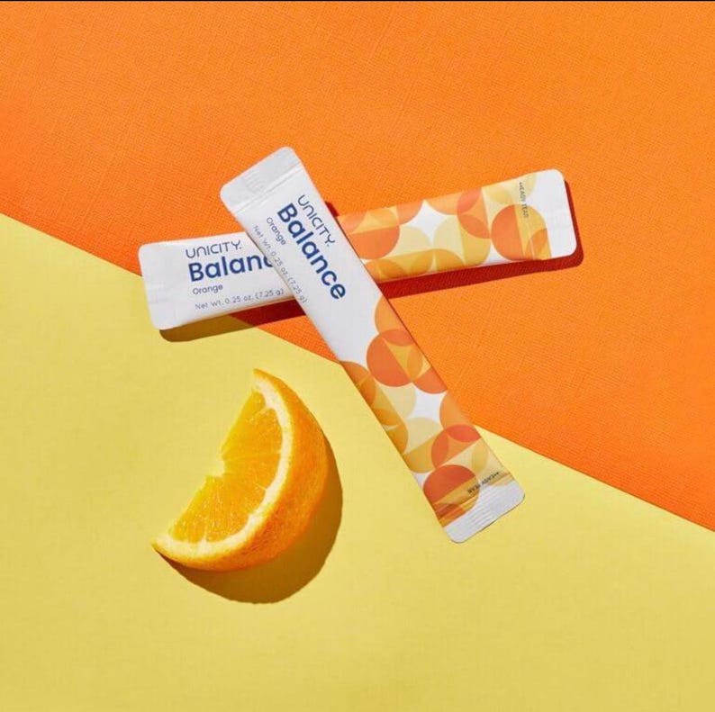 May include: Two white Unicity Balance Orange supplement packets with a geometric orange and yellow circle pattern. Each packet displays the text Unicity Balance Orange and a net weight of 0.25 oz. A fresh orange wedge sits beside the packets.