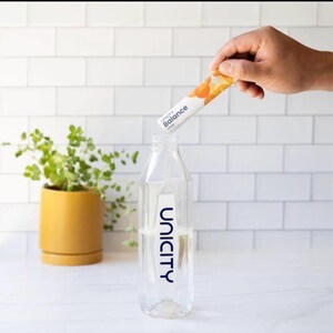 May include: A clear plastic water bottle with the Unicity logo printed in blue on the front. A hand holds a white Unicity Balance packet with orange circular graphics above the bottle opening, ready to pour the contents into the water.