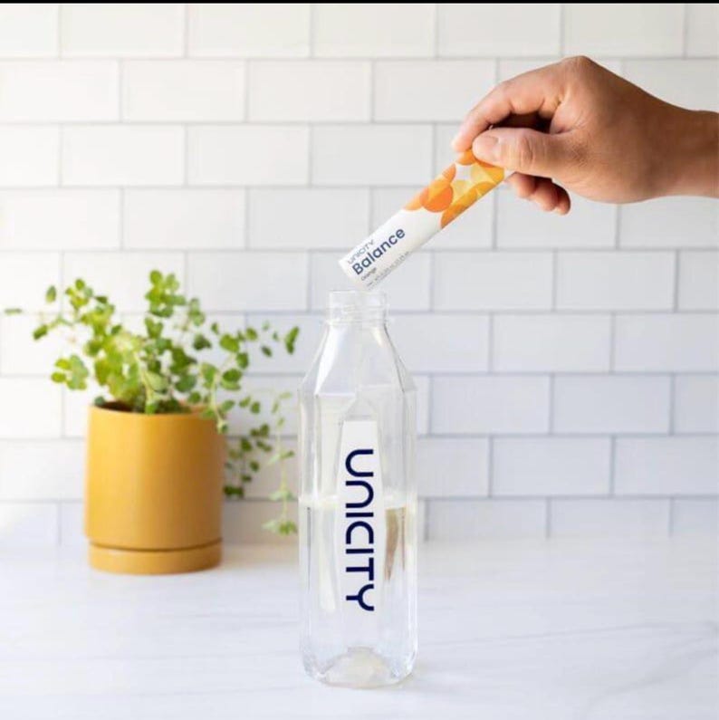 May include: A clear plastic water bottle with the Unicity logo printed in blue on the front. A hand holds a white Unicity Balance packet with orange circular graphics above the bottle opening, ready to pour the contents into the water.