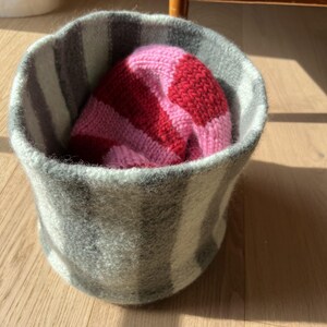 Könnte beinhalten: Ein zylindrischer Filzkorb mit einem vertikalen Streifenmuster in Grau und Weiß. Im Inneren des Korbs liegt eine weiche Strickmütze mit breiten horizontalen Streifen in abwechselnd leuchtendem Pink und tiefem Rot.
