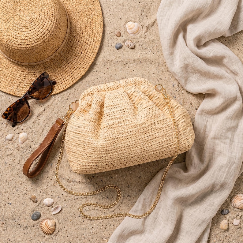 May include: A beach-themed still life featuring a woven straw clutch with a gold chain strap and a brown leather handle. A straw hat and tortoiseshell sunglasses are next to the clutch. A linen scarf and seashells complete the scene.