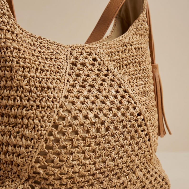 May include: A woven straw shoulder bag with a tan leather strap and a decorative tassel. The bag features a textured, open weave design in a natural beige color, perfect for carrying essentials.