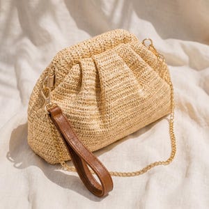 May include: A light beige woven straw clutch bag with a gathered top. It has a brown leather wrist strap, a gold chain strap, and gold-tone hardware. The bag is a neutral colour.