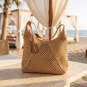 May include: A woven straw tote bag with a brown leather handle and a matching tassel. The bag has a geometric pattern and is sitting on a stone surface. The background shows a beach scene with white draped cabanas and the ocean.