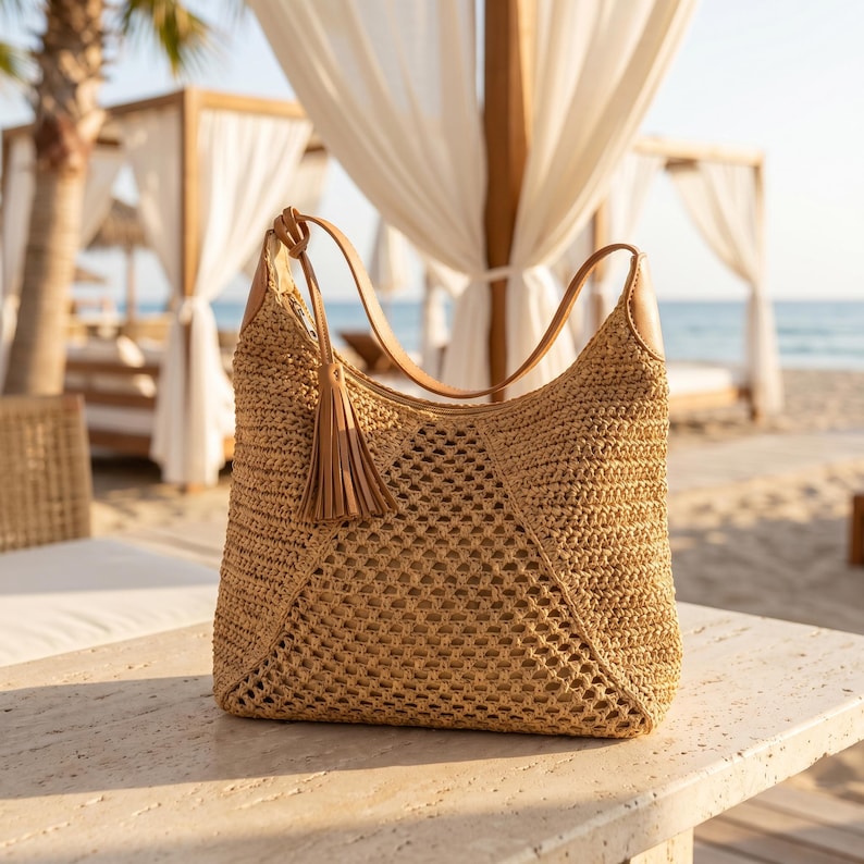 May include: A woven straw tote bag with a brown leather handle and a matching tassel. The bag has a geometric pattern and is sitting on a stone surface. The background shows a beach scene with white draped cabanas and the ocean.