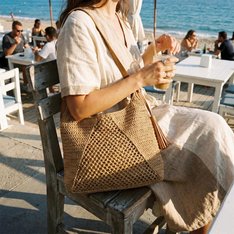 May include: A woven straw tote bag with a geometric design and a leather strap. The bag has a tassel and is tan in color. The person is sitting on a wooden chair near the beach.