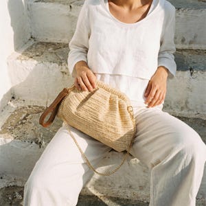 May include: A beige woven clutch bag with a brown leather handle and a gold chain strap. The bag is gathered at the top and held by a person wearing a white top and trousers. The background is white stone steps.