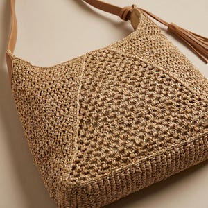 May include: A woven straw shoulder bag with a tan leather strap and tassel. The bag features a geometric design on the front and a neutral color palette. The strap is adjustable and the bag is lightweight.