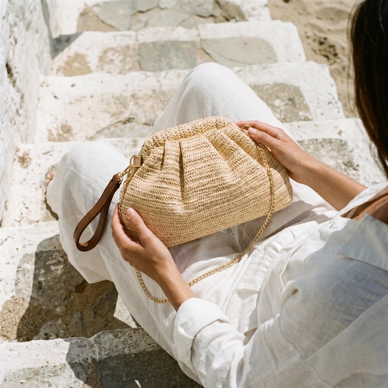 May include: A beige woven clutch bag with a gold chain strap and a brown leather handle. The bag has a gathered design and is held by a person wearing white clothing. The background shows stone steps and a beach.