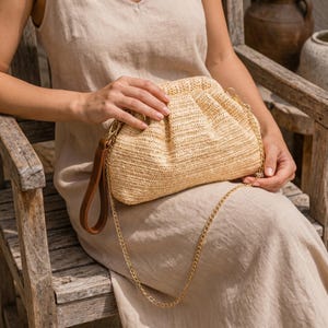 May include: A woven straw clutch bag with a gold chain strap and a brown leather handle. The bag is a light tan colour and has a gathered top. The chain is draped down the front of the bag.