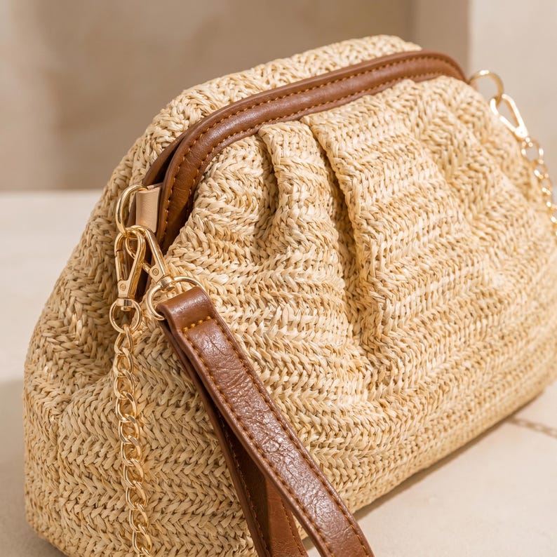May include: A woven straw clutch bag with brown leather trim and a wrist strap. It features a gold chain strap and gold-coloured hardware. The bag is light tan and has a gathered design.