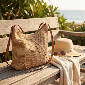 May include: A woven straw tote bag with brown leather straps sits on a wooden bench. A straw hat with a brown band and sunglasses rest beside a cream-colored scarf. The scene is set outdoors with a view of the ocean.