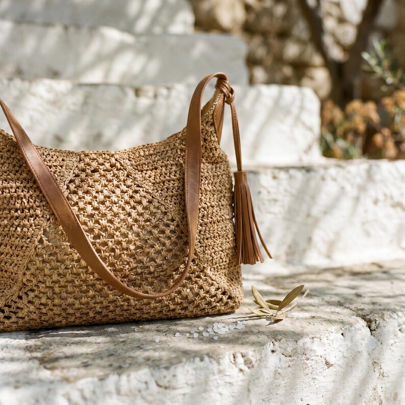 May include: A woven straw shoulder bag with brown leather straps and a tassel. The bag is a light tan color and has a textured, open weave design. The bag is sitting on a stone surface.