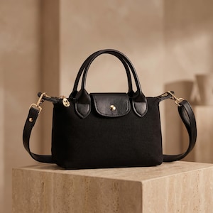 May include: A black handbag featuring a structured canvas body, two short top handles, and a detachable shoulder strap. The bag includes a front flap with a central gold-tone snap button closure and gold-tone metal hardware on the strap attachments.