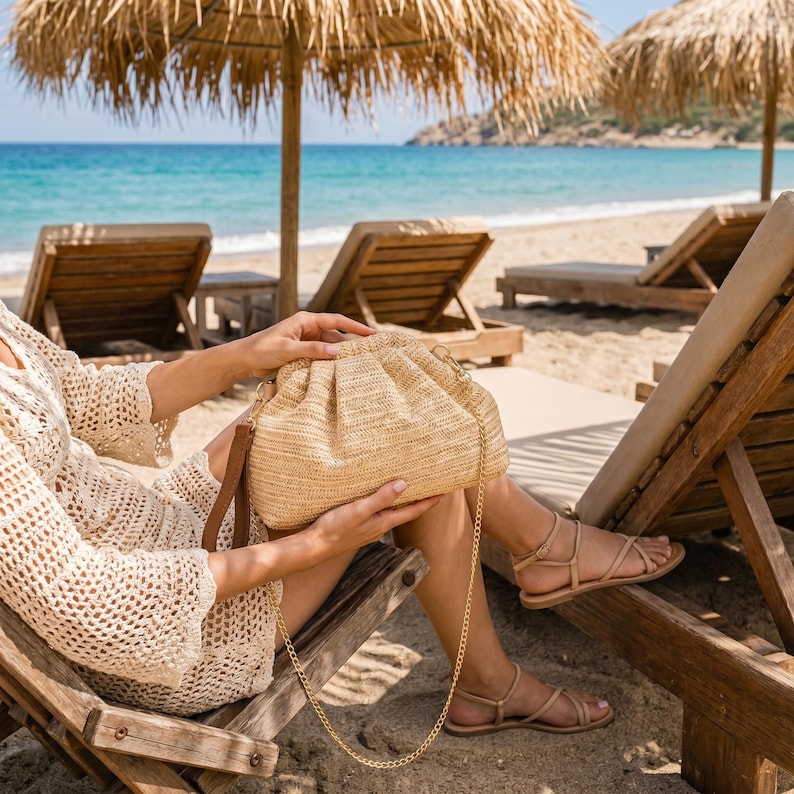 May include: A beige woven handbag with a gold chain strap and brown leather details. The bag is held by a person sitting on a wooden beach chair. The background includes a beach, ocean, and straw parasols.