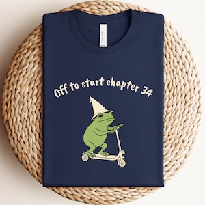May include: A navy blue t-shirt featuring a graphic of a green frog wearing a pointed hat and riding a white scooter. The text above the frog reads "Off to start chapter 34" in a light-colored, rounded font. The shirt has a crew neckline.