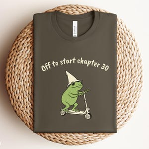 May include: An army green t-shirt featuring a graphic of a green frog wearing a white pointed hat and riding a white scooter. Above the frog, the text reads "Off to start chapter 30" in a light cream color. The shirt has a crew neckline.