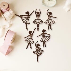 Op de afbeelding: Zes bruine houten ballerina-uitsparingen in verschillende dansposities. Elke uitsparing toont een ballerina in een tutu, met uitgestrekte armen en benen. De uitsparingen zijn gerangschikt op een wit oppervlak, met spoelen lint en een klok op de achtergrond.