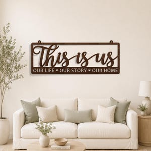 以下が含まれることがあります： 筆記体で「This is us」と書かれた茶色の木製サイン。テキストの下には、ハートのシンボルが付いた「OUR LIFE OUR STORY OUR HOME」という言葉があります。サインは長方形で、白いソファの上に吊り下げられています。