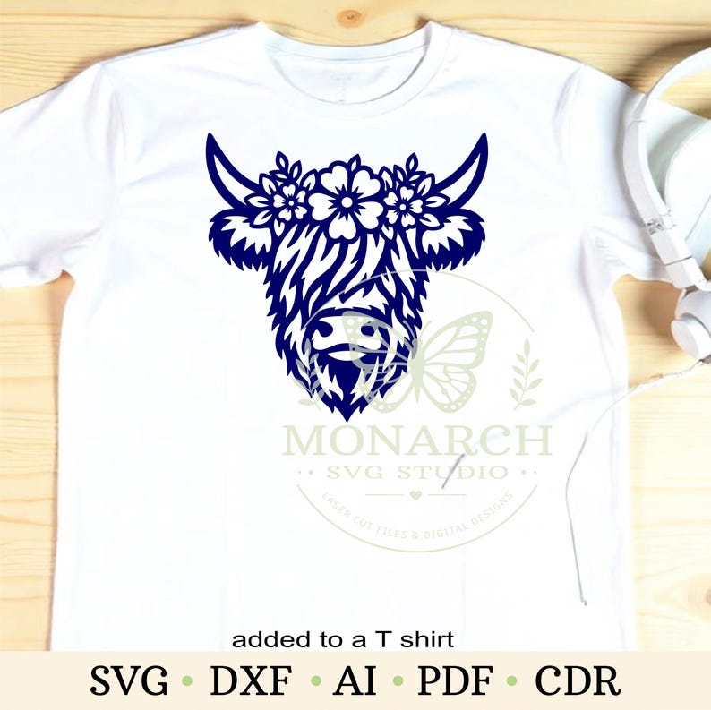 Puede incluir: Una camiseta blanca con un gr&aacute;fico azul oscuro de una cabeza de vaca Highland adornada con flores entre los cuernos. El dise&ntilde;o incluye una marca de agua con un icono de mariposa y el texto MONARCH SVG STUDIO, adem&aacute;s del texto added to a T shirt.