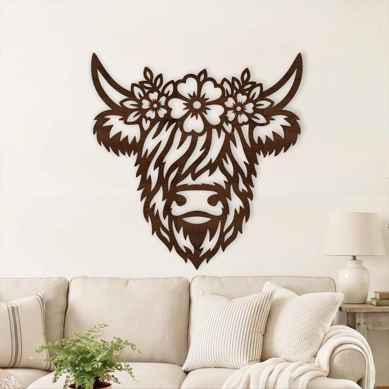Puede incluir: Una pieza de arte mural de madera marr&oacute;n oscuro con forma de cabeza de vaca Highland. El dise&ntilde;o presenta una corona floral entre los cuernos, con intrincados detalles recortados que forman el pelaje peludo, los rasgos faciales y los p&eacute;talos.
