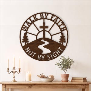 Op de afbeelding: Een rond wandbord van donkerbruin hout met een uitgesneden ontwerp van een kruis op een heuvel met een kronkelend pad, twee dennenbomen en een zon. De tekst "WALK BY FAITH" staat in een boog bovenaan en "NOT BY SIGHT" onderaan.