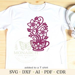 Op de afbeelding: Een wit t-shirt met korte mouwen en een donker magenta afbeelding van een theekopje gevuld met gestileerde bloemen en krullende ranken. Het ontwerp bevat een watermerk met een vlindericoon en de tekst Monarch SVG Studio.