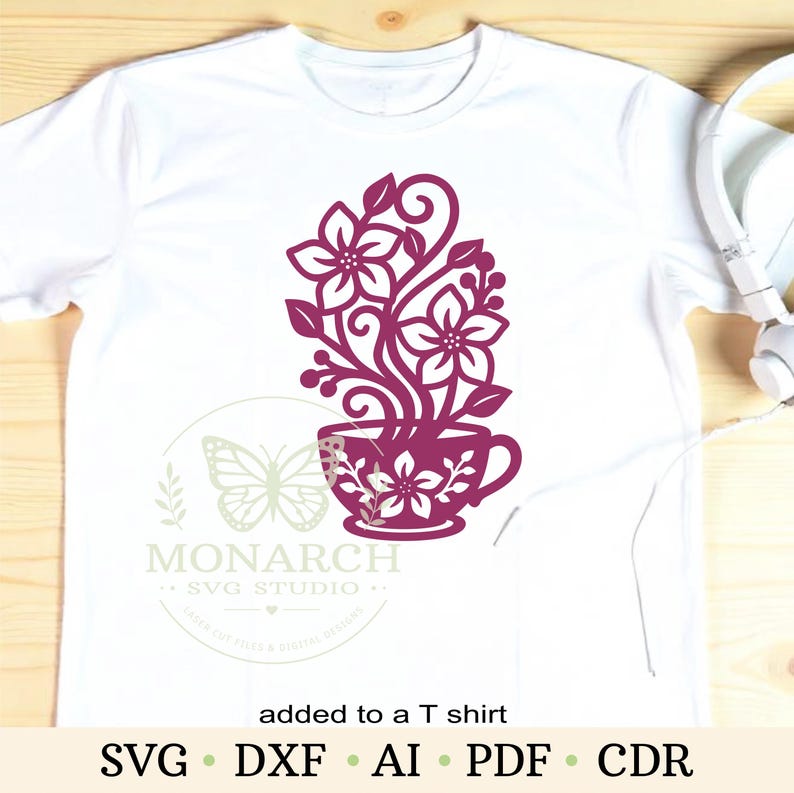 Op de afbeelding: Een wit t-shirt met korte mouwen en een donker magenta afbeelding van een theekopje gevuld met gestileerde bloemen en krullende ranken. Het ontwerp bevat een watermerk met een vlindericoon en de tekst Monarch SVG Studio.