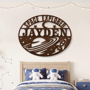 May include: A circular dark brown wooden wall sign featuring the text SPACE EXPLORER and the name JAYDEN in large capital letters. The design includes a crescent moon, several stars, a planet with rings, and a rocket ship silhouette cutout.
