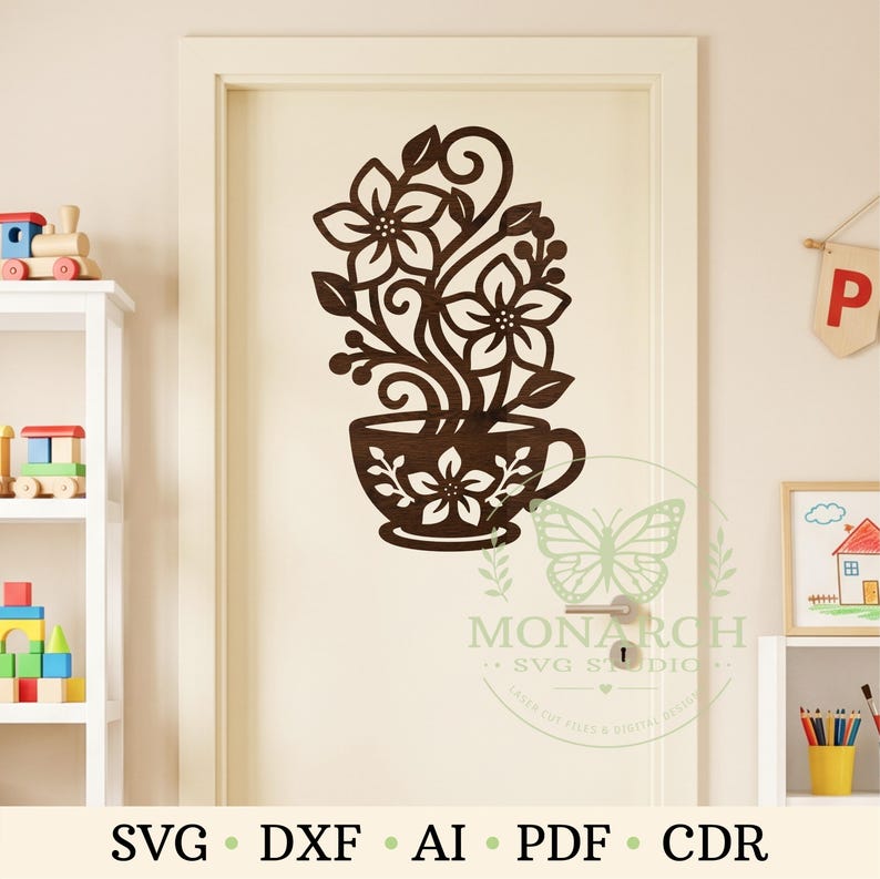 Op de afbeelding: Een donkerbruine, lasergesneden muursticker met een theekopje gevuld met gestileerde bloemen en krullende ranken. Het ontwerp is op een deur aangebracht. Een watermerk met een vlinderpictogram en de tekst MONARCH SVG STUDIO is zichtbaar.