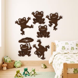 May include: A set of six dark brown wooden frog wall decals in various poses, including one holding a lily pad leaf. The frogs feature cut-out eyes and mouths, displaying a natural wood grain texture, arranged on a plain wall above a bed.