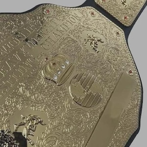 Puede incluir: Un cintur&oacute;n de campeonato de lucha libre chapado en oro con intrincados patrones de filigrana, un emblema central de corona con piedras claras incrustadas y figuras de luchadores en relieve. El texto dice World Heavyweight Wrestling Champion.