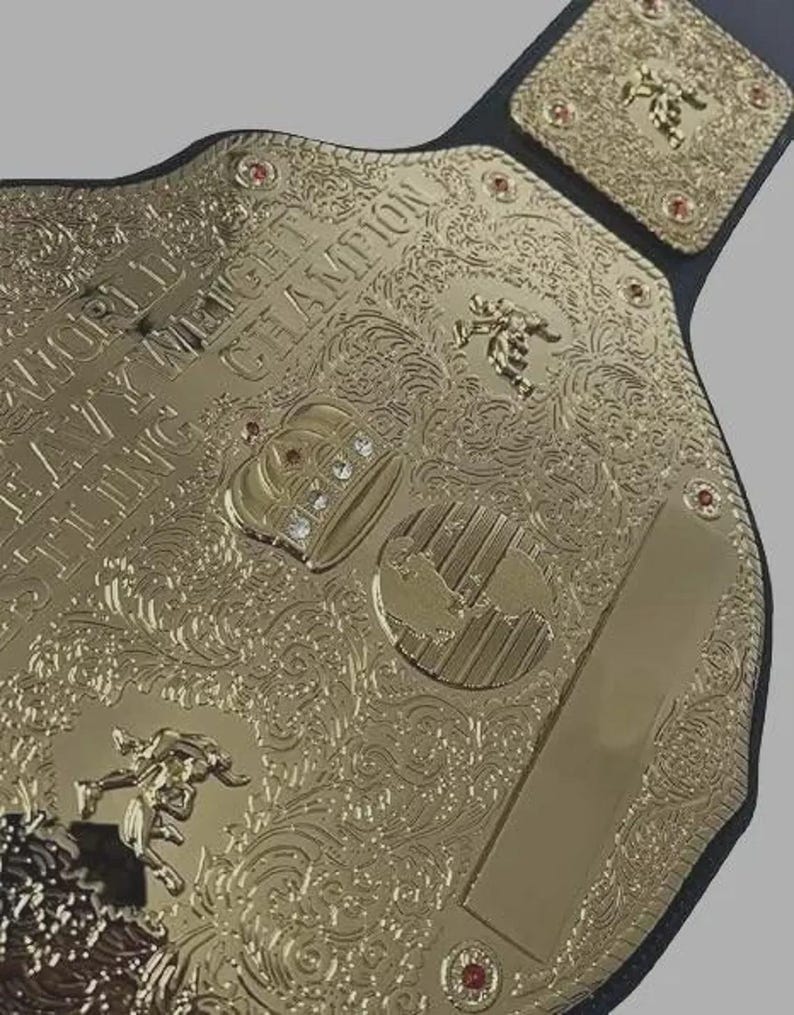 Puede incluir: Un cintur&oacute;n de campeonato de lucha libre chapado en oro con intrincados patrones de filigrana, un emblema central de corona con piedras claras incrustadas y figuras de luchadores en relieve. El texto dice World Heavyweight Wrestling Champion.