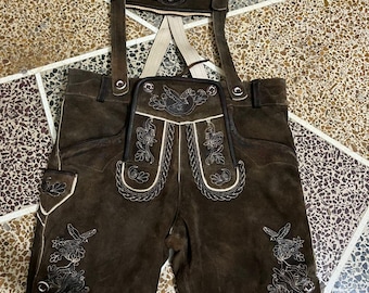 Puede incluir: Lederhosen de cuero ante marrón oscuro con intrincados bordados de colores claros en la solapa frontal, las piernas y el puente del pecho. La prenda incluye tirantes ajustables con hebillas de metal y ribetes blancos a lo largo de las costuras.
