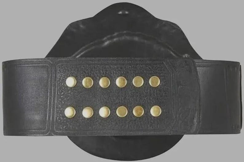 Puede incluir: Un cintur&oacute;n de cuero negro que presenta una secci&oacute;n rectangular con dos filas paralelas de seis peque&ntilde;os remaches met&aacute;licos circulares en tono lat&oacute;n. El cintur&oacute;n tiene un acabado de superficie texturizado y un detalle de borde liso.